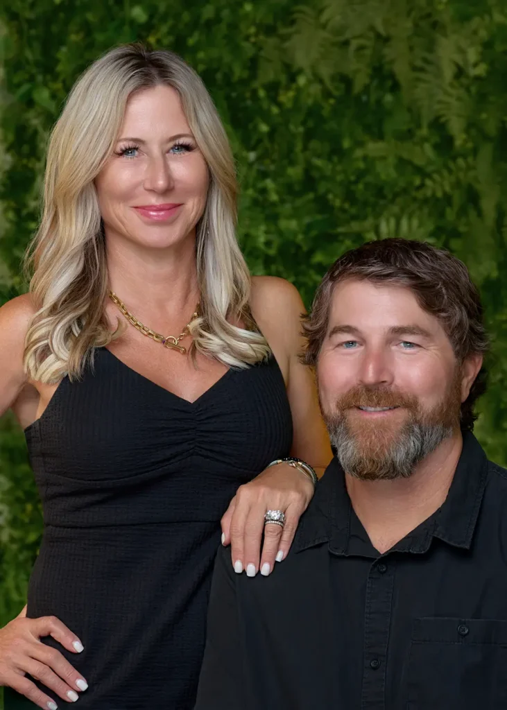 A woman with long blonde hair and a black dress stands beside a seated man with brown hair and a beard, both posing in front of a green leafy background. - The Look in Gladstone and Clinton, NJ