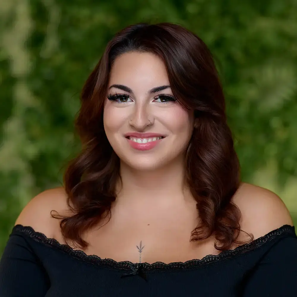 Woman with long brown hair wearing an off-the-shoulder black top, smiling, with a green leafy background. - The Look in Gladstone and Clinton, NJ