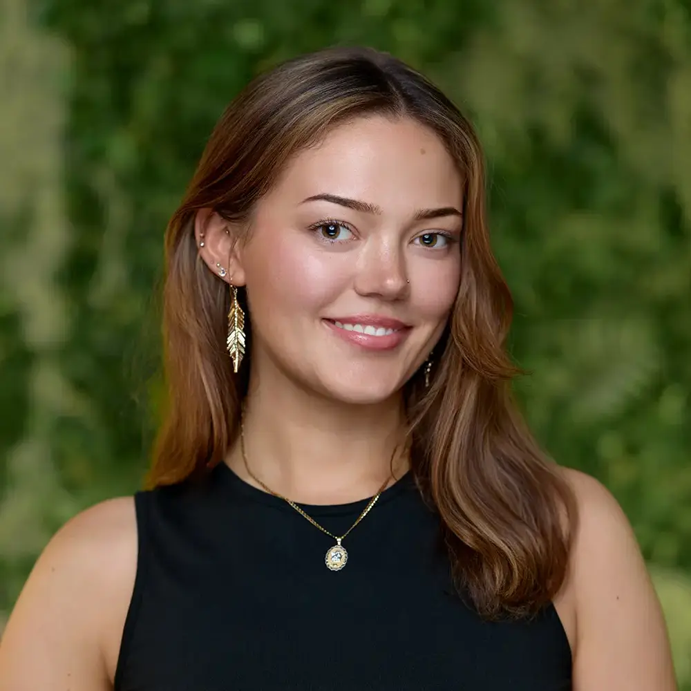 A woman with light brown hair, wearing a black sleeveless top, gold earrings, and a necklace, stands in front of a green, leafy background and smiles at the camera. - The Look in Gladstone and Clinton, NJ